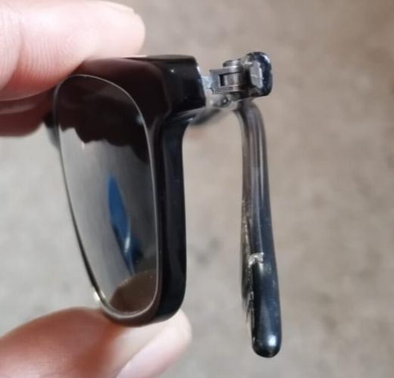 May include: Close-up of black sunglasses with a rectangular frame. The sunglasses have a dark lens and a silver hinge. The arm of the sunglasses is also black. The sunglasses are partially open.