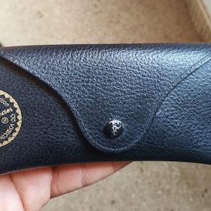 May include: A dark blue, textured sunglasses case with a snap closure. The case features a gold-colored Ray-Ban logo and text on the front. The case is designed to protect eyewear.