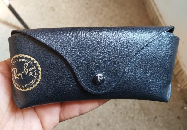 May include: A dark blue, textured sunglasses case with a snap closure. The case features a gold-colored Ray-Ban logo and text on the front. The case is designed to protect eyewear.