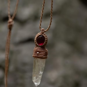 May include: A macrame pendant necklace featuring a clear, pointed crystal point suspended from a tan cord. The crystal is secured by a tan woven macrame setting that includes a small, smooth, dark red oval stone at the top and a tiny silver bead.