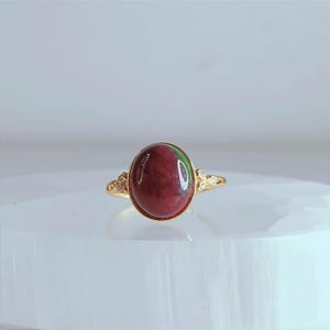 May include: A gold ring with an oval, deep red gemstone with green hues. The ring has a detailed design with small, clear stones on either side of the gemstone. The ring is set against a soft, white background. The ring is a piece of jewellery.