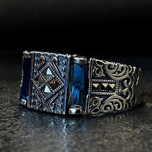 Könnte beinhalten: Ein silberner Ring mit aufwendigem Filigran und rechteckigen blauen Edelsteinen. Der Ring hat einen zentralen blauen Stein, der von kleineren Steinen und dekorativen Mustern flankiert wird. Das Design des Rings ist symmetrisch und detailliert.