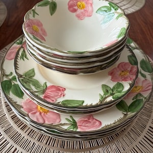 May include: A set of cream-coloured ceramic plates and bowls with a floral design. The plates and bowls feature pink roses with yellow centres and green leaves. The rims are trimmed with a brown border. The set is arranged on a woven placemat.