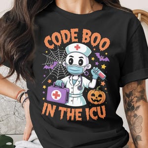 Può includere: T-shirt nera con grafica a tema Halloween. Il design presenta un'infermiera dei cartoni animati con una maschera, che tiene una siringa e una borsa medica, con la scritta "CODE BOO IN THE ICU". Una zucca, pipistrelli e ragnatele completano il design.