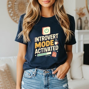 May include: Navy blue t-shirt with the text "INTROVERT MODE ACTIVATED" in yellow and white. The design includes a power button, coffee cup, and a "DO NOT DISTURB" shopping bag icon. The shirt is paired with blue jeans.