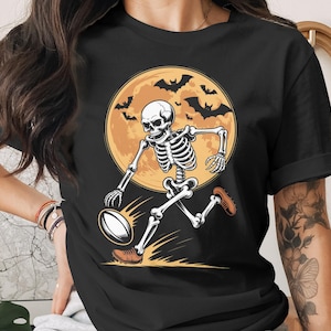 May include: Black t-shirt featuring a skeleton playing rugby in front of a full moon with bats. The skeleton is wearing brown shoes and the rugby ball is white and brown. The design is a Halloween theme.