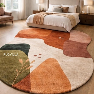 Op de afbeelding: Een ovaal vloerkleed met een abstract ontwerp in een slaapkamer. Het vloerkleed heeft een kleurenpalet van oranje, bruin, groen en crème. Het vloerkleed heeft een modern design. De merknaam "RUGVILLA" staat op het vloerkleed.