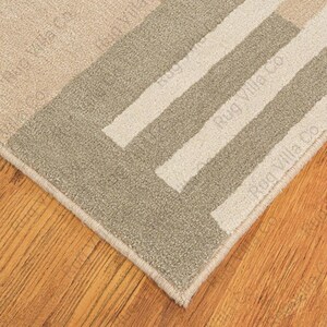 K&ouml;nnte beinhalten: Nahaufnahme eines beigefarbenen und grauen Teppichs mit geometrischem Muster. Der Teppich hat ein rechteckiges Design mit Streifen und einem Rand. Der Text "Rug Villa Co" ist auf dem Teppich aufgedruckt. Der Teppich liegt auf einem Holzboden.