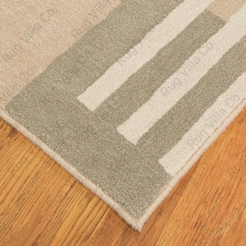 K&ouml;nnte beinhalten: Nahaufnahme eines beigefarbenen und grauen Teppichs mit geometrischem Muster. Der Teppich hat ein rechteckiges Design mit Streifen und einem Rand. Der Text "Rug Villa Co" ist auf dem Teppich aufgedruckt. Der Teppich liegt auf einem Holzboden.