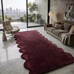 May include: A deep burgundy rug with a distinctive wavy edge, set in a contemporary living space. The rug sits on a marble floor, with a city view visible through a large window. A sofa and chair are also present.