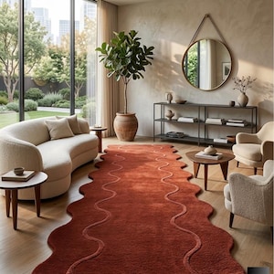 May include: A terracotta-coloured rug with a wavy edge and a central, meandering design. The rug is placed in a living room setting with a curved cream-coloured sofa, two armchairs, and a large window overlooking a garden.
