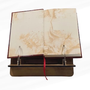 May include: An open book with cream pages and gold marbling, displayed on a wooden stand. The book has a red ribbon bookmark and is held open by clear acrylic supports. The stand is made of wood.
