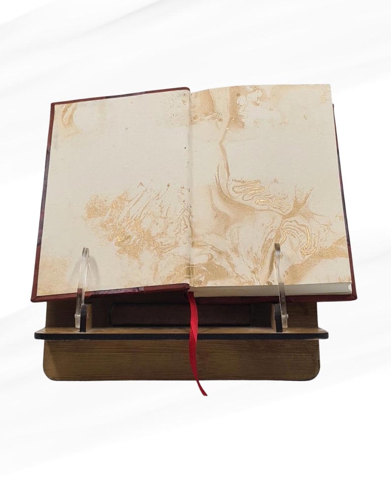 May include: An open book with cream pages and gold marbling, displayed on a wooden stand. The book has a red ribbon bookmark and is held open by clear acrylic supports. The stand is made of wood.
