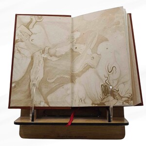 Peut inclure: Un livre ouvert avec des pages marbr&eacute;es, pr&eacute;sent&eacute; sur un pr&eacute;sentoir en bois avec un signet en ruban rouge. Le livre a une couverture rouge fonc&eacute; et les pages ont un motif beige et marron. Le pr&eacute;sentoir est en bois et poss&egrave;de un support en acrylique transparent.