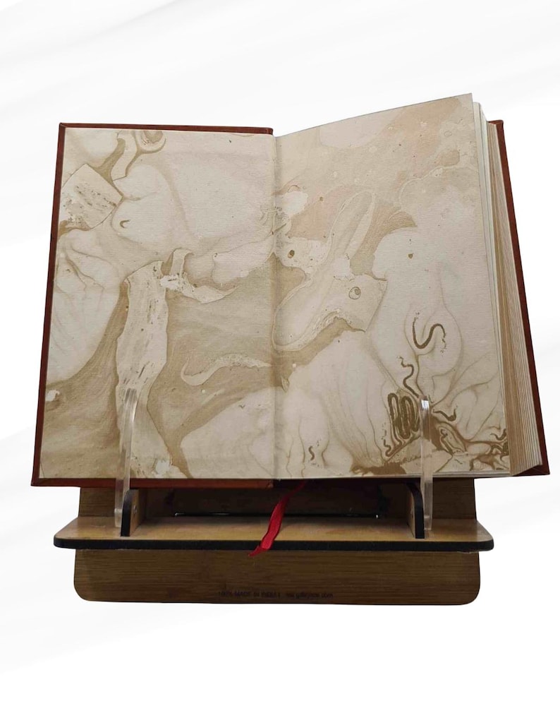 Peut inclure: Un livre ouvert avec des pages marbr&eacute;es, pr&eacute;sent&eacute; sur un pr&eacute;sentoir en bois avec un signet en ruban rouge. Le livre a une couverture rouge fonc&eacute; et les pages ont un motif beige et marron. Le pr&eacute;sentoir est en bois et poss&egrave;de un support en acrylique transparent.