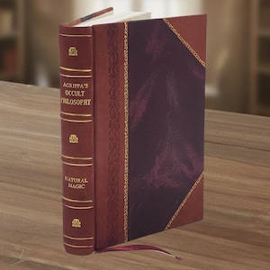 Puede incluir: Un libro de tapa dura titulado "AGRIPPA'S OCCULT PHILOSOPHY NATURAL MAGIC" con una cubierta de cuero burdeos y detalles dorados. El libro tiene un panel morado oscuro en la portada y una cinta de marcador burdeos. El libro est&aacute; de pie sobre una superficie de madera.