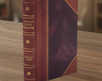 Op de afbeelding: Een hardcover boek getiteld "AGRIPPA'S OCCULT PHILOSOPHY NATURAL MAGIC" met een bordeauxrode leren kaft en gouden accenten. Het boek heeft een donkerpaars paneel op de voorkant en een bordeauxrood lint als bladwijzer. Het boek staat rechtop op een houten oppervlak.