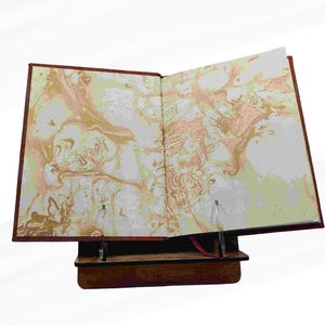 Puede incluir: Un libro abierto con p&aacute;ginas veteadas, expuesto sobre un atril de madera. La cubierta del libro es de color rojo intenso y las p&aacute;ginas presentan un dise&ntilde;o arremolinado de oro, melocot&oacute;n y blanco. El atril es de color marr&oacute;n oscuro.