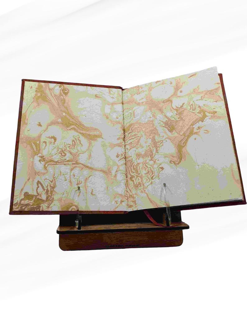 Puede incluir: Un libro abierto con p&aacute;ginas veteadas, expuesto sobre un atril de madera. La cubierta del libro es de color rojo intenso y las p&aacute;ginas presentan un dise&ntilde;o arremolinado de oro, melocot&oacute;n y blanco. El atril es de color marr&oacute;n oscuro.