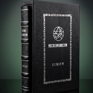 Puede incluir: Un libro de tapa dura negro titulado Necronomicon de Simon, con texto plateado en relieve y un sello circular en la portada. El lomo tiene nervios con detalles plateados y la cubierta est&aacute; bordeada por un intrincado patr&oacute;n en relieve.