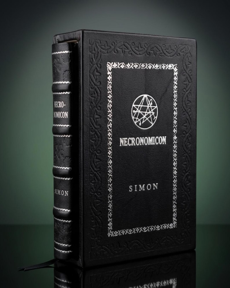 Puede incluir: Un libro de tapa dura negro titulado Necronomicon de Simon, con texto plateado en relieve y un sello circular en la portada. El lomo tiene nervios con detalles plateados y la cubierta est&aacute; bordeada por un intrincado patr&oacute;n en relieve.