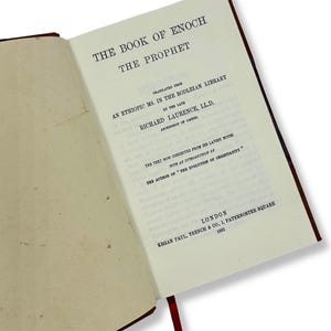May include: An open book titled "THE BOOK OF ENOCH THE PROPHET" with text in black font. The book's pages are off-white, and the cover is a dark reddish-brown. A red ribbon bookmark is visible. The text includes the translator's name and the publisher's location.
