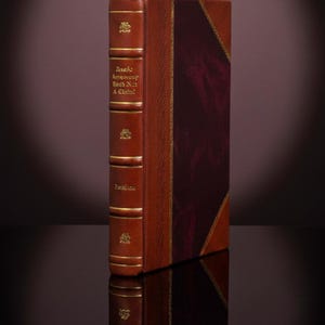 May include: A hardback book with a leather cover. The spine is a lighter shade of brown with gold lettering and decorative elements. The cover has a burgundy panel with gold trim. The book is reflected in a dark surface.