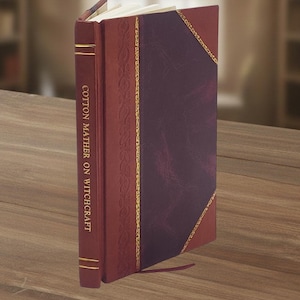 May include: A hardcover book titled "Cotton Mather on Witchcraft" with a burgundy leather cover and gold accents. The book has a decorative spine and a dark purple center panel. A red ribbon bookmark is visible.