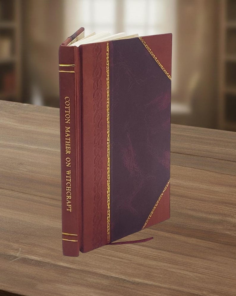 May include: A hardcover book titled "Cotton Mather on Witchcraft" with a burgundy leather cover and gold accents. The book has a decorative spine and a dark purple center panel. A red ribbon bookmark is visible.