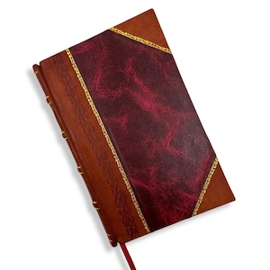 May include: A leather-bound journal with a burgundy cover and a brown spine. The cover features a textured burgundy center panel framed by gold trim. A red ribbon bookmark is visible.