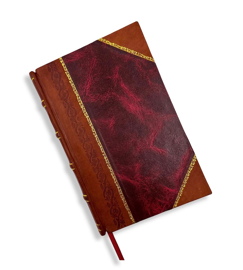 May include: A leather-bound journal with a burgundy cover and a brown spine. The cover features a textured burgundy center panel framed by gold trim. A red ribbon bookmark is visible.