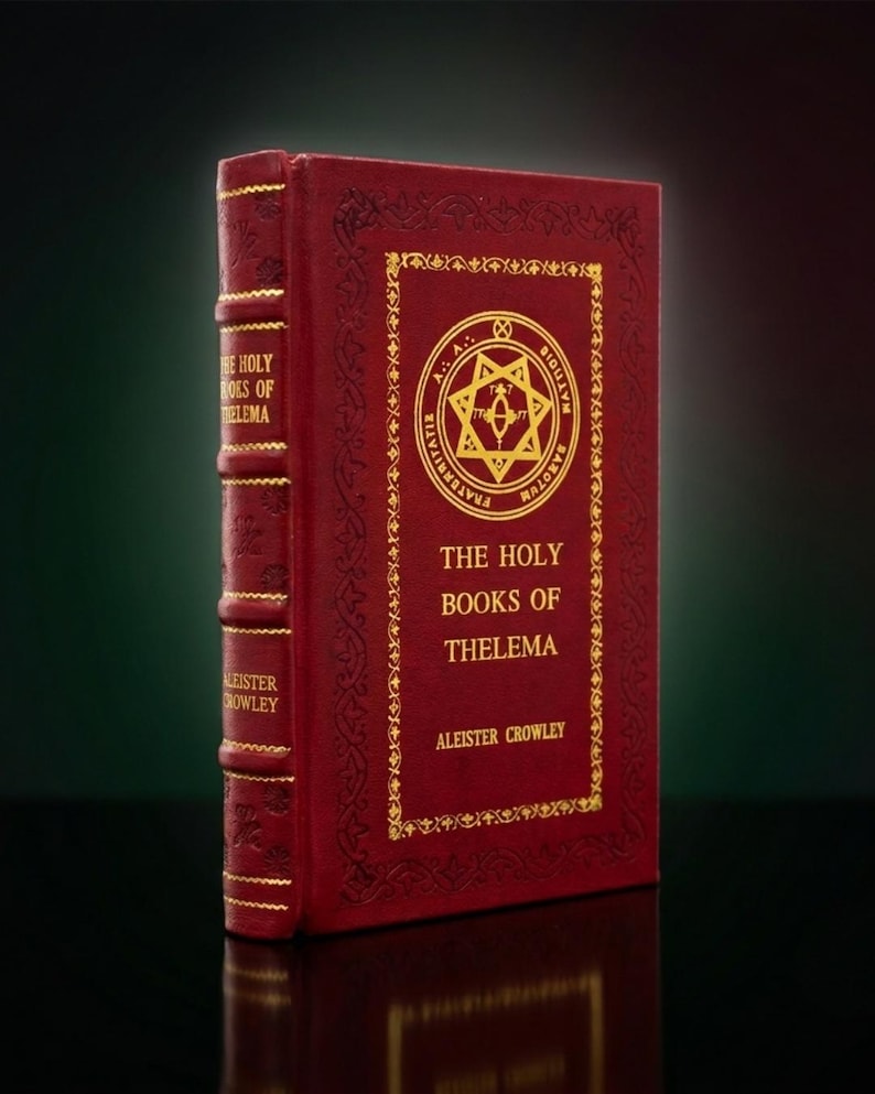 Op de afbeelding: Een donkerrood gebonden boek getiteld The Holy Books of Thelema door Aleister Crowley. De voorkant heeft een goudkleurig reli&euml;f van een cirkelvormig embleem met een ster en tekst, omringd door een decoratieve rand. De rug heeft gouden letters.
