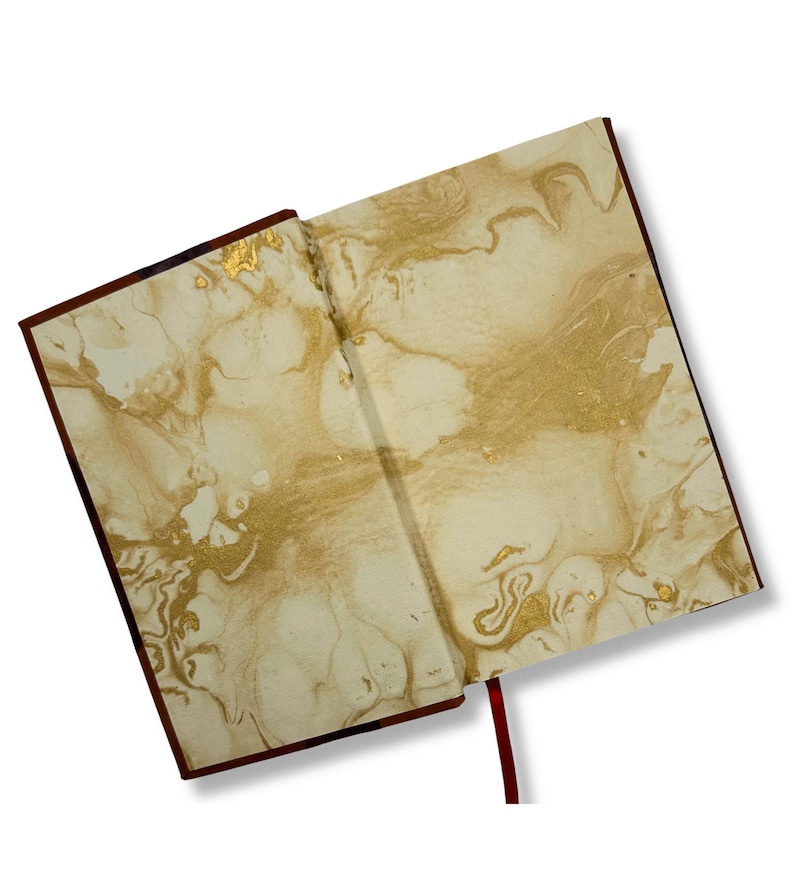 May include: Open journal with a cream-colored marbled design and gold accents. The journal has a dark brown cover and a red ribbon bookmark. The pages are open, displaying the intricate pattern.