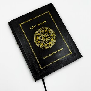 May include: A black book with gold lettering and a gold geometric design. The text "Liber Azerate" is at the top, with "Magister Templi Frater Nemidial" at the bottom. A black ribbon bookmark is visible.