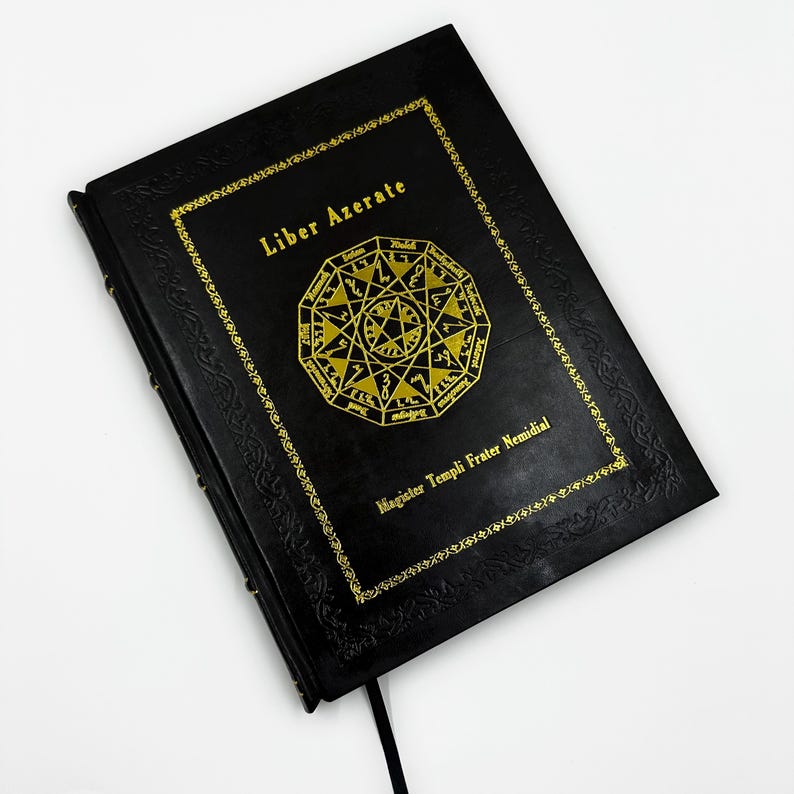 May include: A black book with gold lettering and a gold geometric design. The text "Liber Azerate" is at the top, with "Magister Templi Frater Nemidial" at the bottom. A black ribbon bookmark is visible.