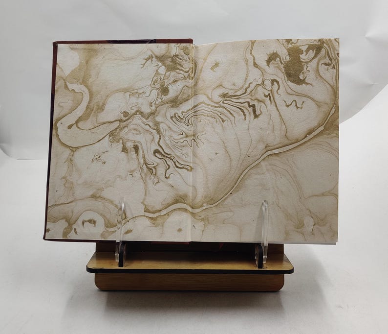 May include: An open book with marbled pages, displayed on a wooden stand. The pages feature a swirling pattern in shades of beige and gold. The book's spine is a deep burgundy color, and the stand is made of light-colored wood.