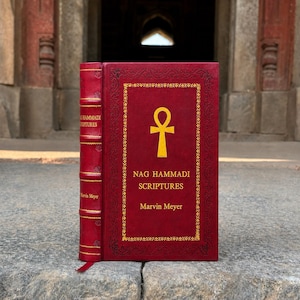 Könnte beinhalten: Ein tiefrotes gebundenes Buch mit dem Titel The Nag Hammadi Scriptures von Marvin Meyer. Das Cover zeigt ein goldenes Ankh-Symbol, goldene Schriftzüge und einen dekorativen Rand. Der Buchrücken hat fünf Bünde mit Goldnähten und Goldtext.