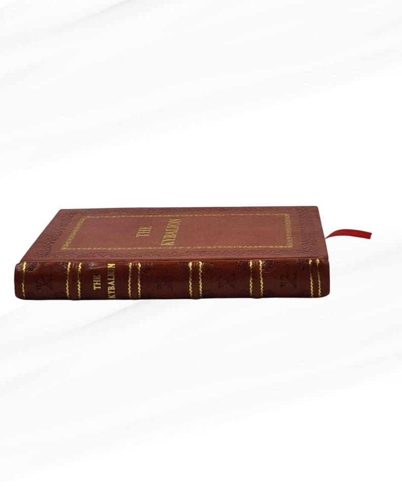 Pode incluir: Um livro de capa dura intitulado "The Kybalion" com capa de couro marrom e letras douradas. O livro tem uma borda decorativa e uma fita vermelha como marcador. A lombada tamb&eacute;m apresenta letras douradas.
