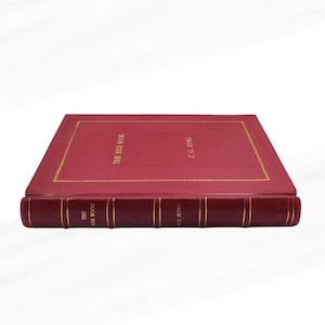 Może przedstawiać: Twarda książka w kolorze czerwonym zatytułowana "THE RED BOOK" autorstwa C.G. Junga. Książka ma złote litery i złotą obw&oacute;dkę. Grzbiet r&oacute;wnież ma złote litery. Książka jest wyświetlana na białym tle.