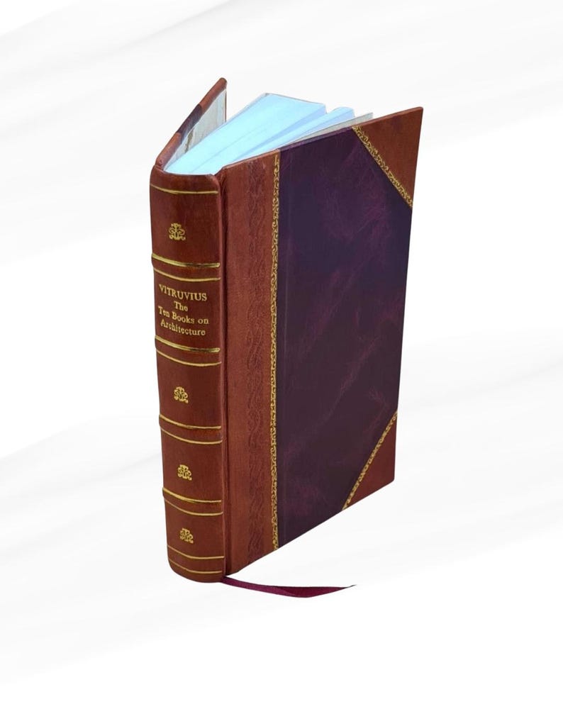 May include: An open book titled "Vitruvius The Ten Books on Architecture." The book has a brown leather cover with gold accents and a burgundy spine. A burgundy ribbon bookmark is visible. The pages are light blue.