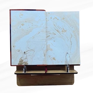 May include: An open book with marbled pages is displayed on a wooden stand. The book's pages have a cream and gold marbled design. The stand is made of light brown wood and has two clear acrylic supports to hold the book open.