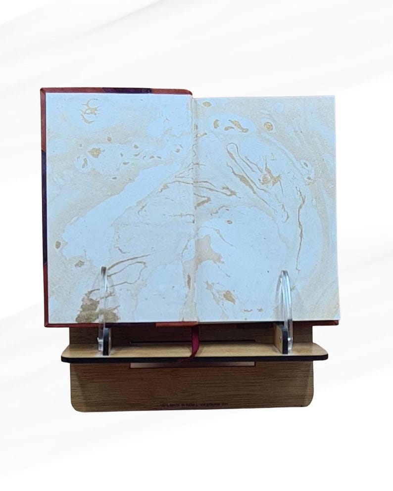 May include: An open book with marbled pages is displayed on a wooden stand. The book's pages have a cream and gold marbled design. The stand is made of light brown wood and has two clear acrylic supports to hold the book open.