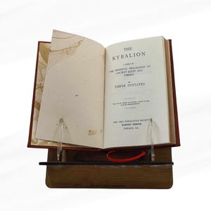 Pode incluir: Um livro aberto intitulado "The Kybalion" &eacute; exibido em um suporte de madeira. O livro tem uma capa vermelha e as p&aacute;ginas s&atilde;o mantidas abertas por suportes de acr&iacute;lico transparente. O t&iacute;tulo e o autor s&atilde;o vis&iacute;veis.