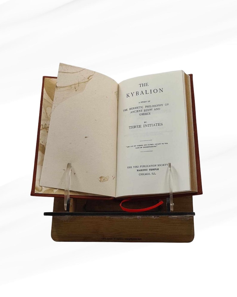 Pode incluir: Um livro aberto intitulado "The Kybalion" &eacute; exibido em um suporte de madeira. O livro tem uma capa vermelha e as p&aacute;ginas s&atilde;o mantidas abertas por suportes de acr&iacute;lico transparente. O t&iacute;tulo e o autor s&atilde;o vis&iacute;veis.