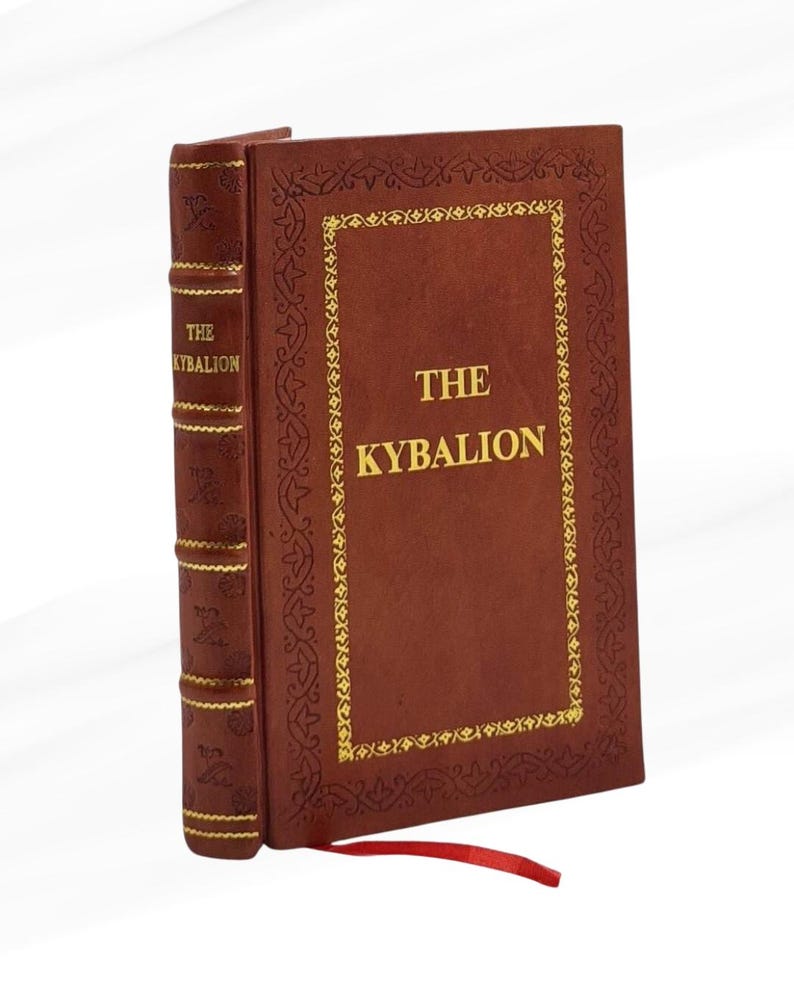Pode incluir: Um livro de capa dura intitulado "THE KYBALION" com capa de couro marrom e letras douradas. A capa apresenta uma borda decorativa dourada. Uma fita vermelha de marcador se estende da lombada do livro.