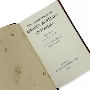 May include: An open book titled "The Meditations of Marcus Aurelius Antoninus" with text in black font. The book is translated by John Jackson and published by Oxford at the Clarendon Press in 1906. The pages are off-white with brown spots.