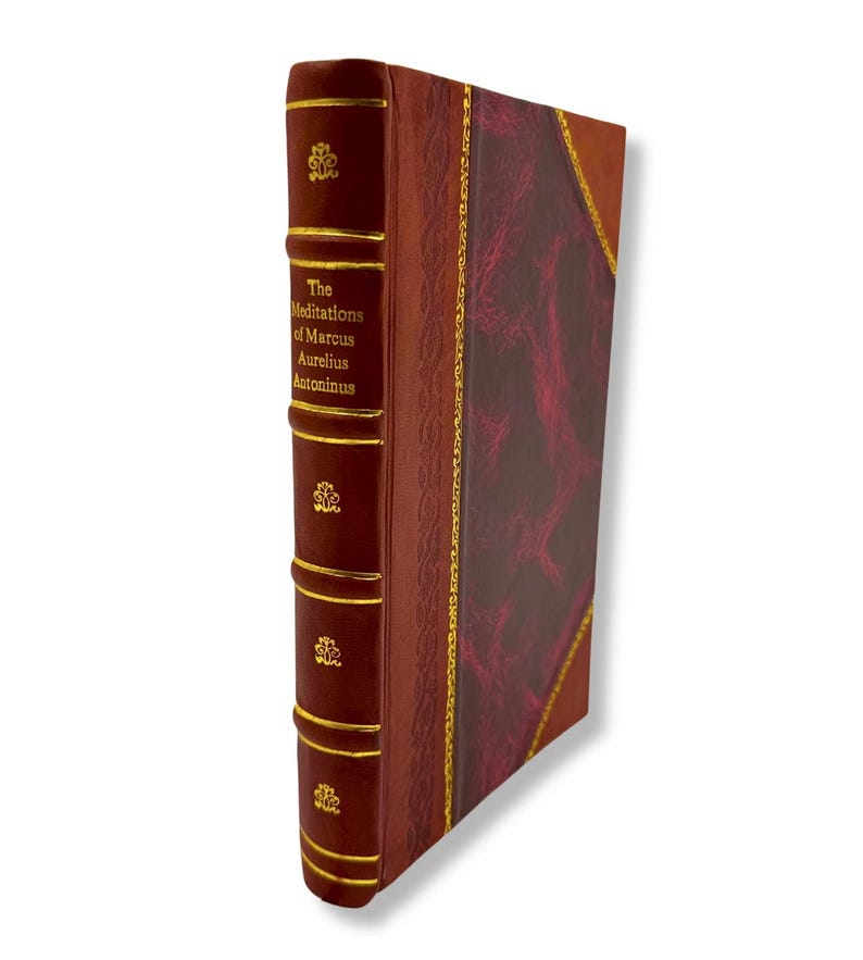 May include: A hardcover book titled "The Meditations of Marcus Aurelius Antoninus." The book has a burgundy leather cover with gold accents and a decorative spine. The edges of the pages are also gold.