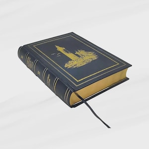 K&ouml;nnte beinhalten: Ein marineblaues Buch mit goldfarbenen Kanten und einer goldenen Leuchtturm-Illustration auf dem Einband. Das Buch hat ein schwarzes Leseband. Der Buchr&uuml;cken hat goldene Schrift.