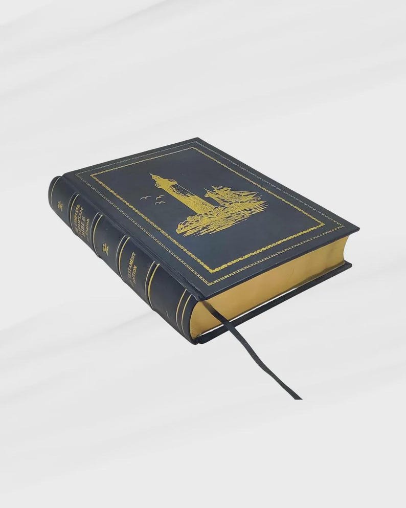 K&ouml;nnte beinhalten: Ein marineblaues Buch mit goldfarbenen Kanten und einer goldenen Leuchtturm-Illustration auf dem Einband. Das Buch hat ein schwarzes Leseband. Der Buchr&uuml;cken hat goldene Schrift.