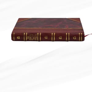 May include: A burgundy leather-bound book with gold detailing. The spine displays the title "THE GOSPEL OF THE HOLY TWELVE" in gold. A red ribbon bookmark is inserted. The book has a classic, elegant design.